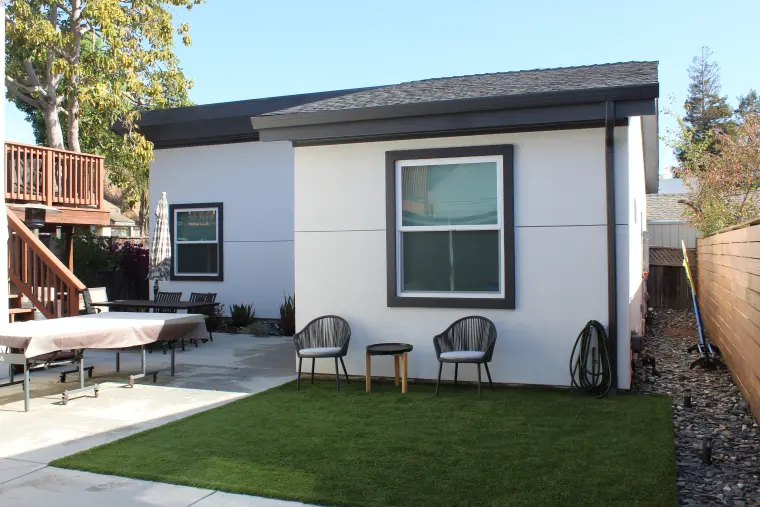 ADU with dark trim and a shed roof, two chairs on artificial turf, and a patio with a dining table beside the main house deck
