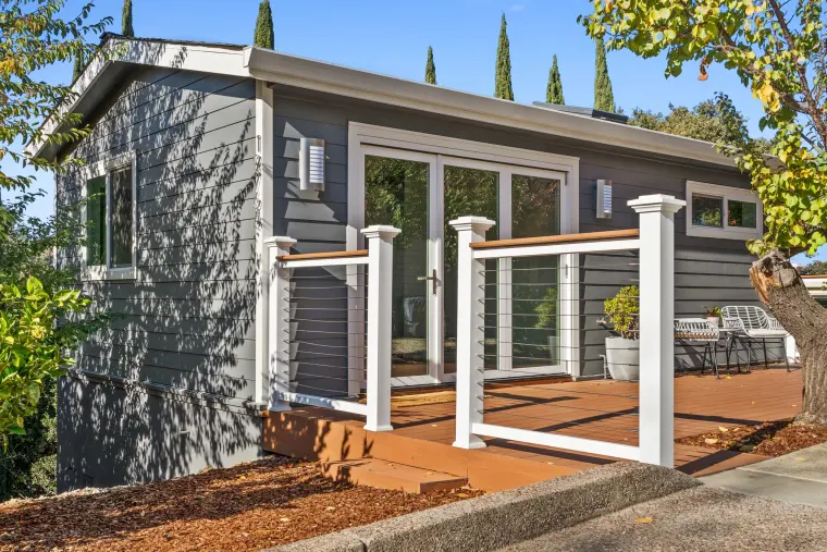 Gray-sided hilltop ADU with a white-railed deck and gate, framed by cypress trees