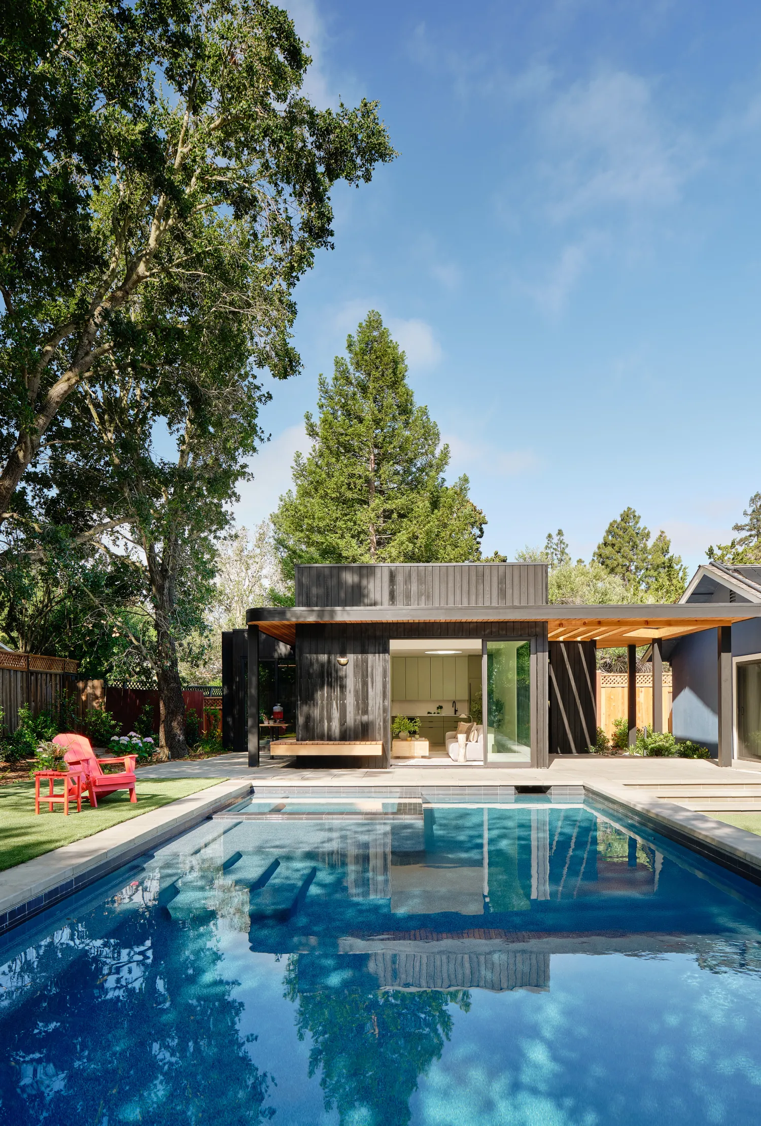 Charred-cedar ADU at the far end of a pool, beneath tall oaks and a redwood, with a red Adirondack chair on the lawn