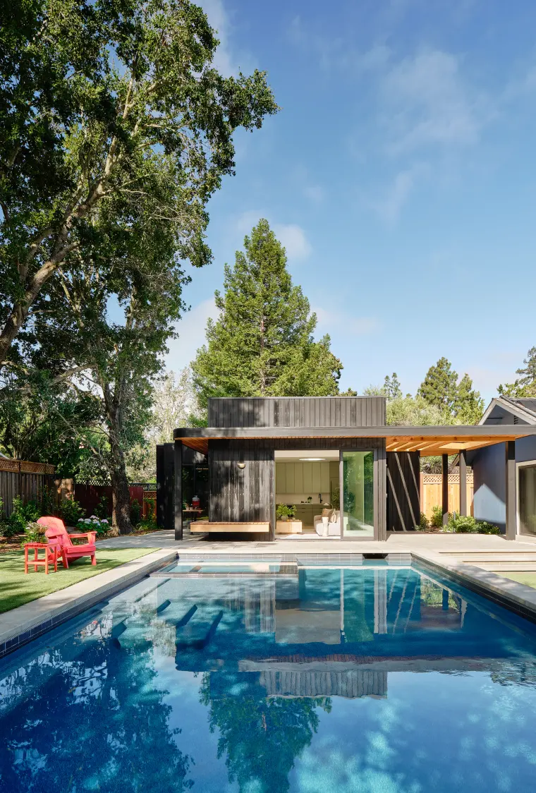 Charred-cedar ADU at the far end of a pool, beneath tall oaks and a redwood, with a red Adirondack chair on the lawn