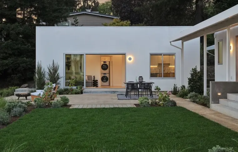 Flat-roofed white stucco ADU at dusk with lit sliding doors, a patio dining set, and a lawn bordered by planting beds