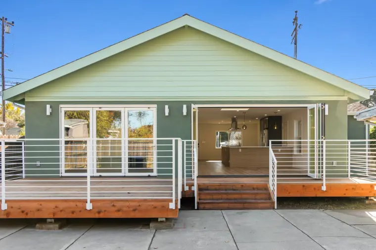 Back of the house with the new addition, a wide wood deck, white metal railings, and glass doors open to the kitchen