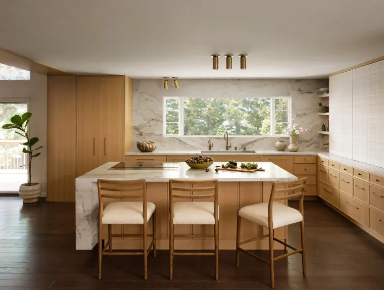 White oak kitchen with a stone-clad island, cushioned counter stools, brass ceiling lights, and a wide window over the sink